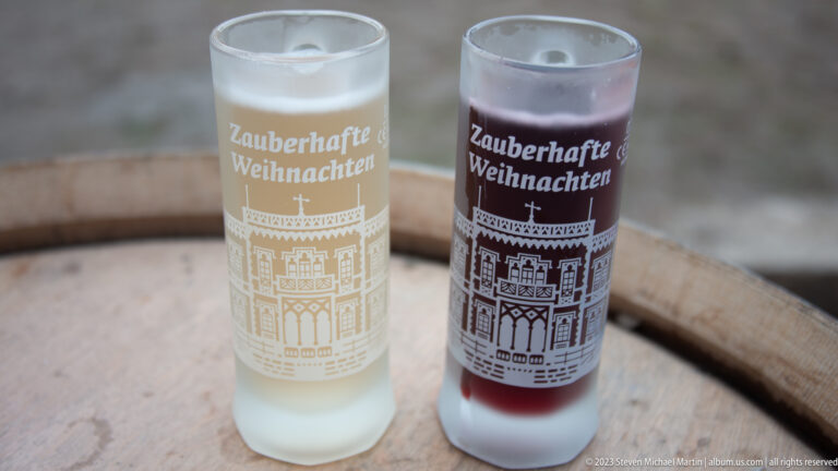 Two frosted Freiburg Christmas glasses on a wooden barrel, holding pale and dark red drinks and printed “Zauberhafte Weihnachten.”