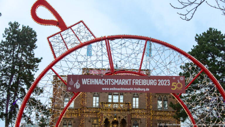 Red illuminated arch labeled Weihnachtsmarkt Freiburg 2023 and 50 frames a light-covered historic building among trees.