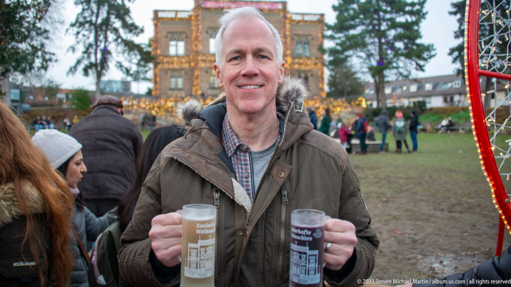 Smiling man holds two tall Christmas-market drinks on a crowded Freiburg lawn, with the red market arch behind him.