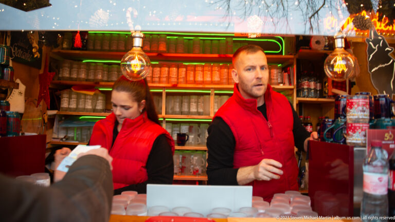 Two vendors in red vests serve customers from a brightly lit Freiburg Christmas-market beverage stall.