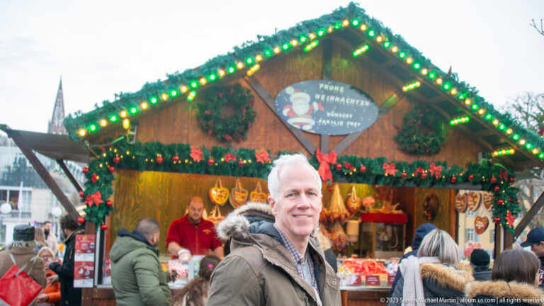 Smiling man stands before a green-lit wooden Christmas-market stall decorated with evergreen garlands as shoppers crowd the counter.