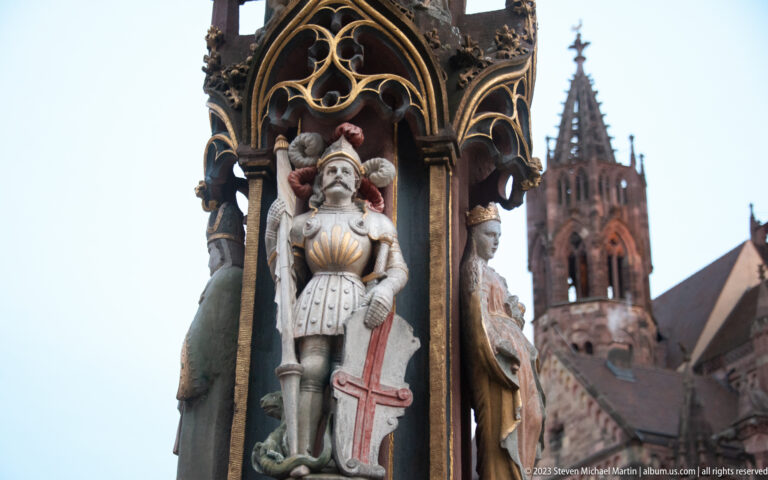 Armored fountain figure beneath an ornate canopy with Freiburg Minster tower blurred in the background.