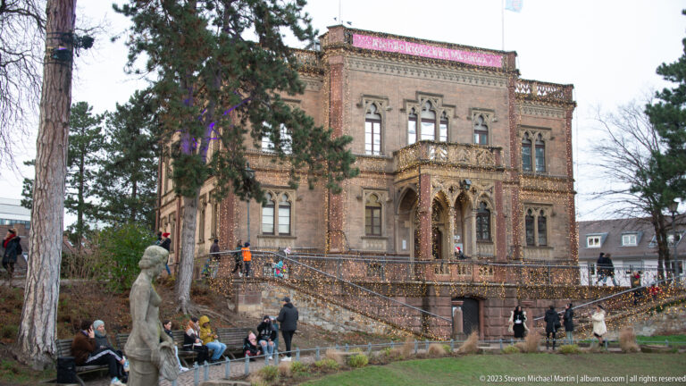 Ornate Colombischlössle with pointed-arch porch, balcony, tall windows, and visitors in the landscaped grounds below.