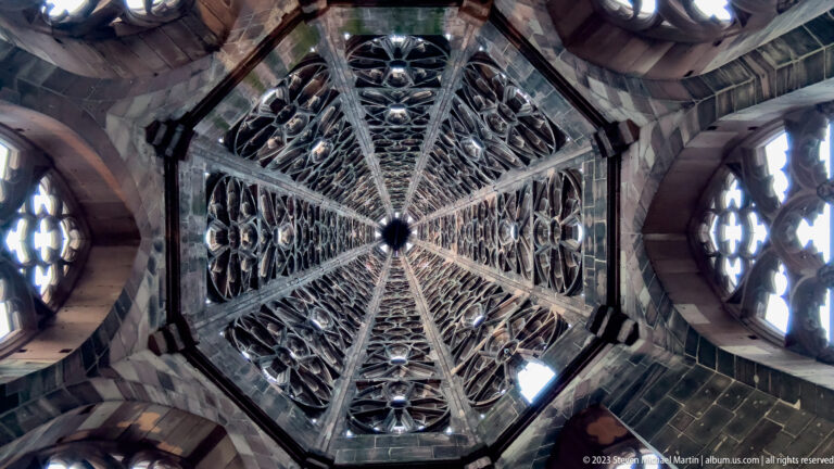Upward view through Freiburg Minster’s Gothic tower, with stone ribs, tracery, and open windows converging overhead.