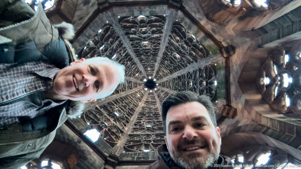 Two visitors smiling beneath the Gothic stone tracery and converging ribs inside Freiburg Minster’s tower.