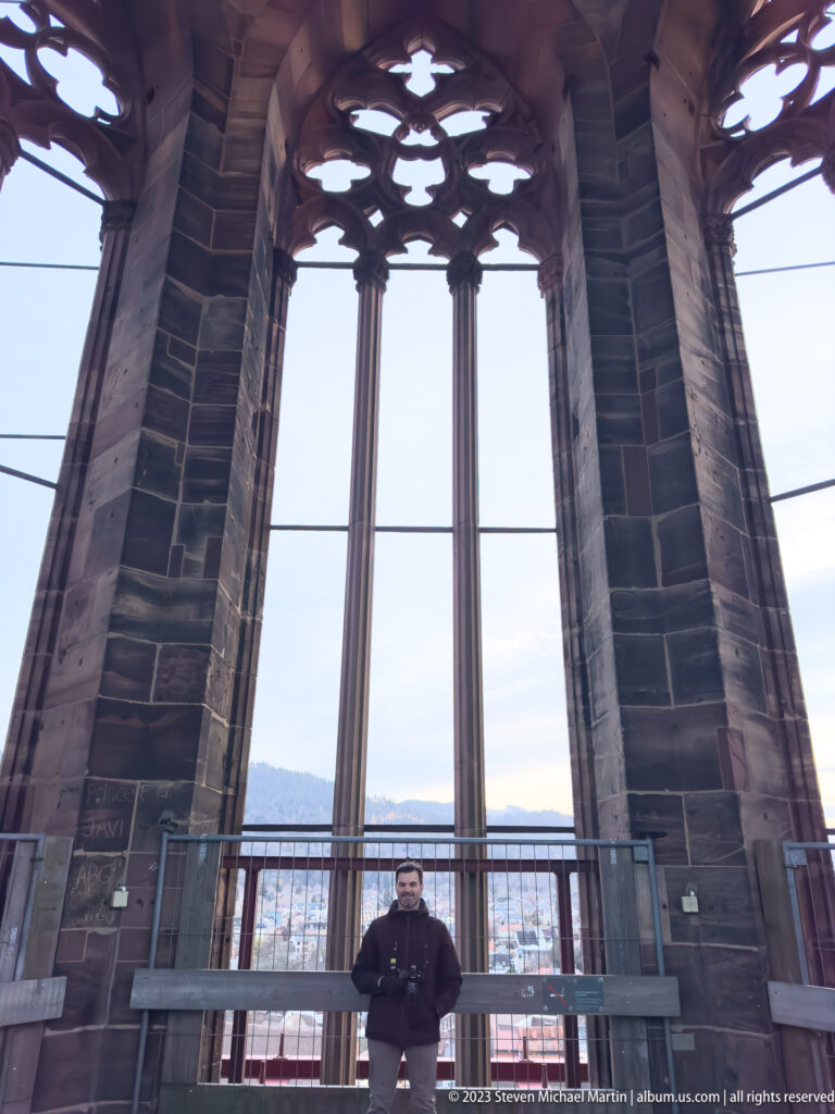 Visitor standing at a tall Gothic opening in Freiburg Minster’s tower, with Freiburg and distant hills visible beyond.