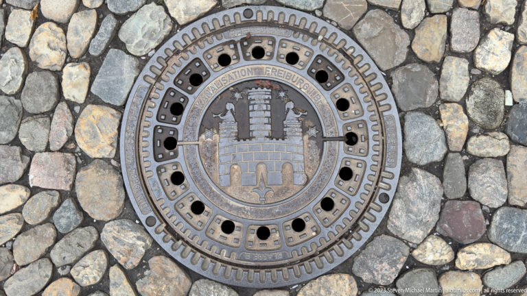 Round Freiburg sewer cover set in cobblestones, bearing the city emblem and the words “KANALISATION FREIBURG.”