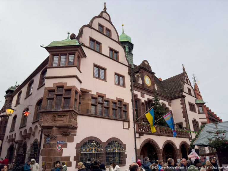 Pale gabled façade of Freiburg’s Neues Rathaus with projecting bays, arched ground floor, clock, flags, and people below.