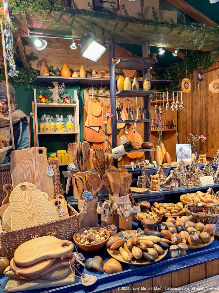 Christmas-market stall filled with wooden bowls, boards, spoons, baskets, and carved crafts beneath evergreen garlands.