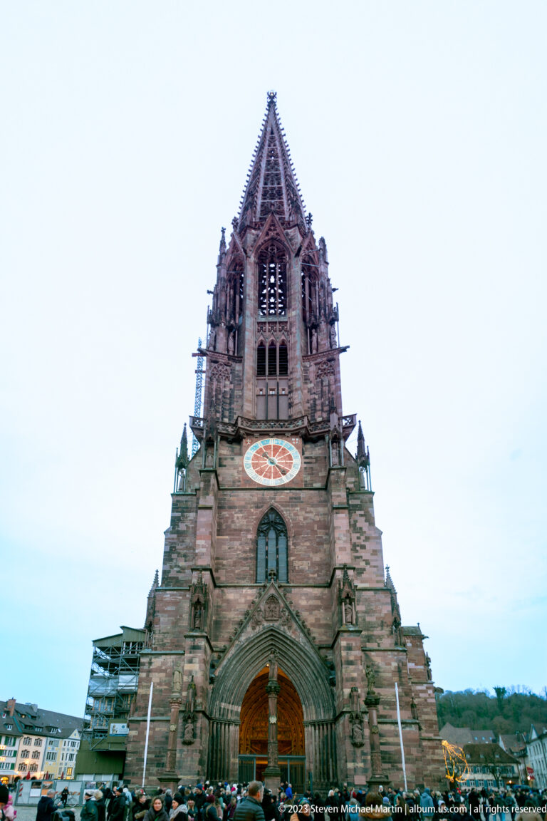 Frontal view of Freiburg Minster west entrance and tall openwork Gothic tower above a crowd in Münsterplatz.