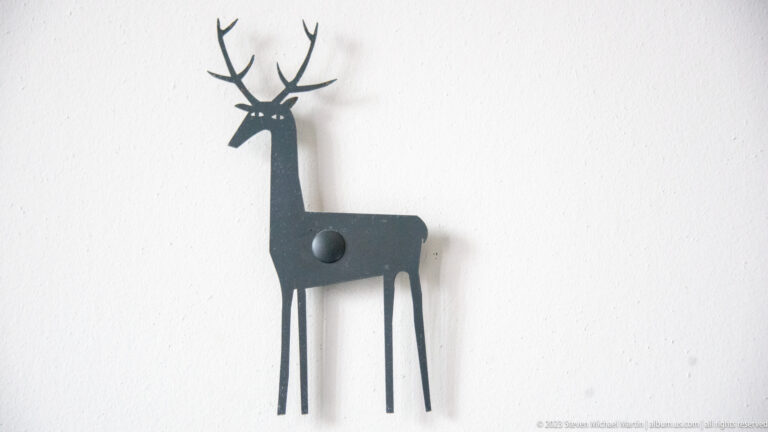 Black metal deer silhouette with branching antlers mounted on a white wall, casting a soft shadow to one side.