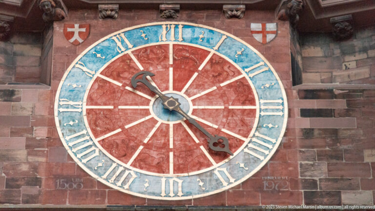 Large red-and-blue clock face with Roman numerals set into the red sandstone tower of Freiburg Minster.