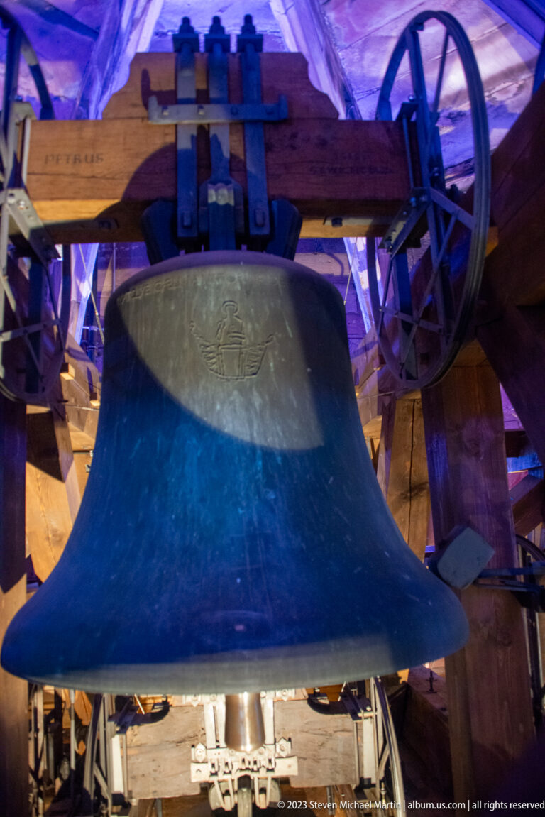 Large bronze cathedral bell suspended from heavy timber beams with wheel mechanism and colored interior lighting.