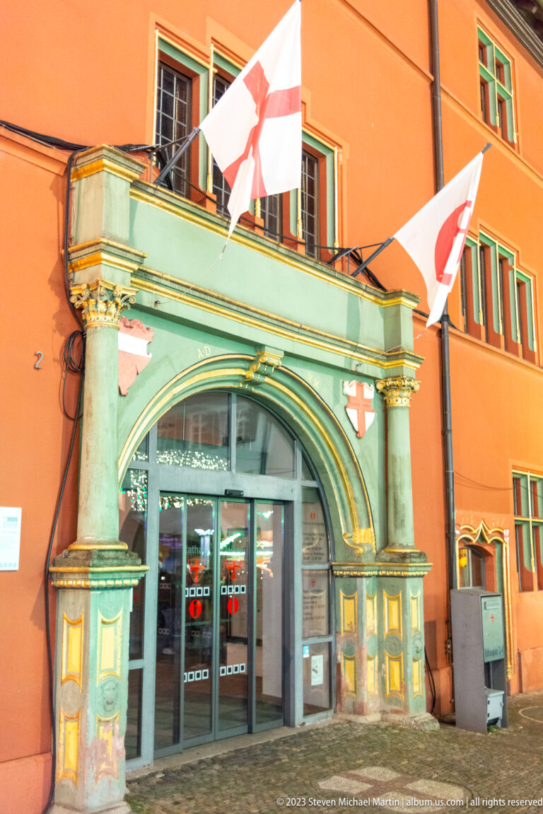 Ornate arched doorway with painted columns and red-and-white flags on a warmly lit historic Freiburg façade.