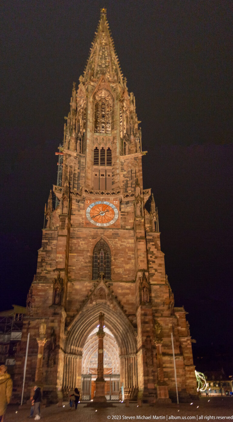 Low-angle nighttime view of Freiburg Minster’s illuminated west façade, portal, clock, and Gothic tower.