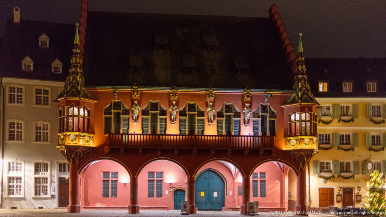Illuminated red Historisches Kaufhaus façade with arcades, balcony, heraldry, and projecting corner oriels.