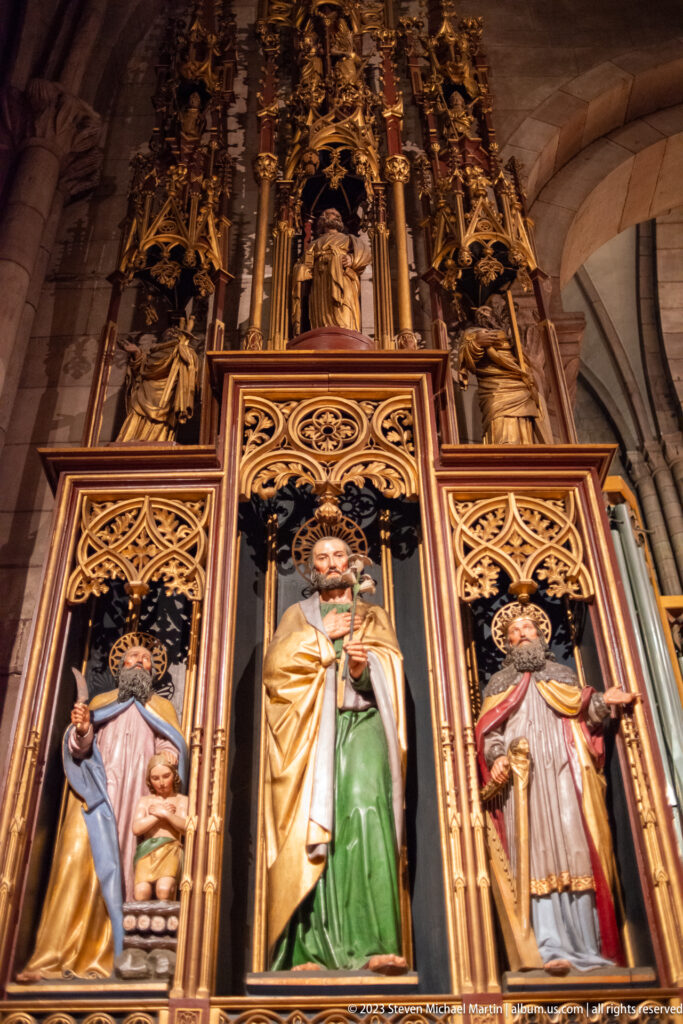 Polychrome sculpted altarpiece with three large figures framed by gilded Gothic tracery inside Freiburg Minster.