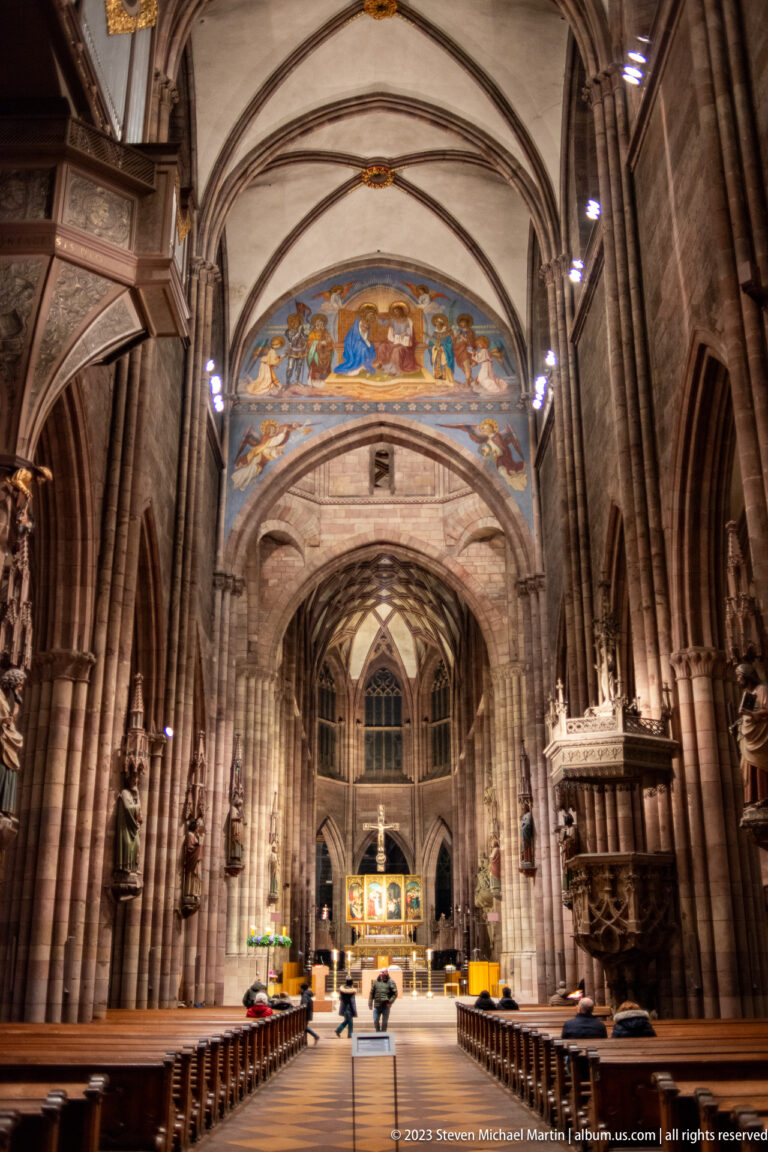 Central nave of Freiburg Minster with pointed arches, ribbed vaults, pews, visitors, and choir beyond.