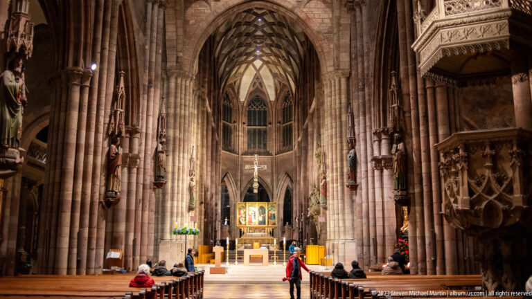 Axial view down Freiburg Minster nave toward the lit altar, with pews, sculptures, and pulpit.