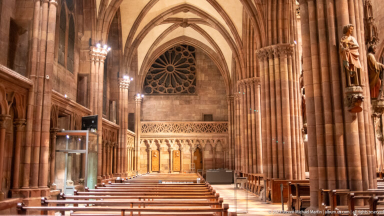 Gothic side aisle in Freiburg Minster with ribbed vaults, wooden seating, sculptures, and circular tracery window.