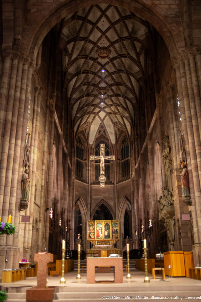 Choir and altar of Freiburg Minster beneath high ribbed Gothic vaults with sculptures and colorful altarpiece.