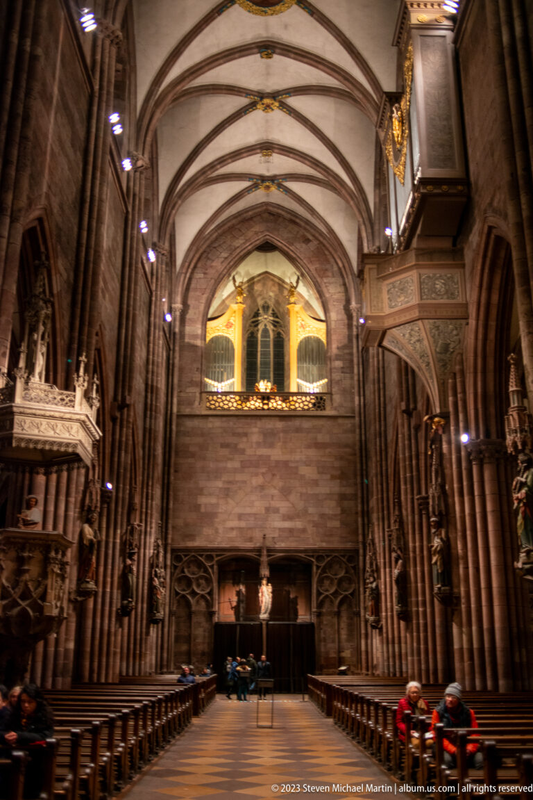 Freiburg Minster nave with ribbed vaults, carved pulpit, seated visitors, and large gallery or organ loft.