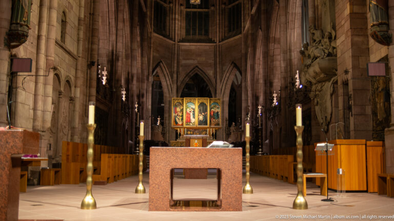 Modern stone altar with tall brass candlesticks and colorful historic altarpiece beyond in Freiburg Minster.