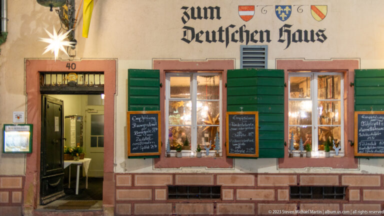 Zum Deutschen Haus façade with green shutters, blackletter sign, lit windows, and chalkboard menus.