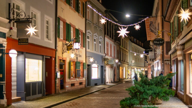 Narrow Freiburg Old Town street with suspended star lights, glowing shopfronts, historic façades, and few pedestrians.