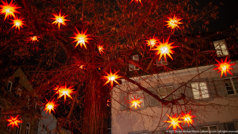 Glowing red star lanterns hanging among leafless tree branches above a Freiburg building at night.