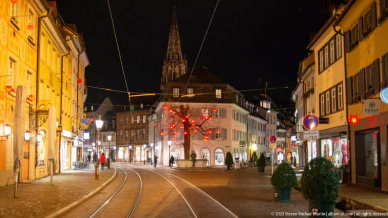 Tram tracks curve through Freiburg toward the distant illuminated spire of Freiburg Minster under Christmas lights.