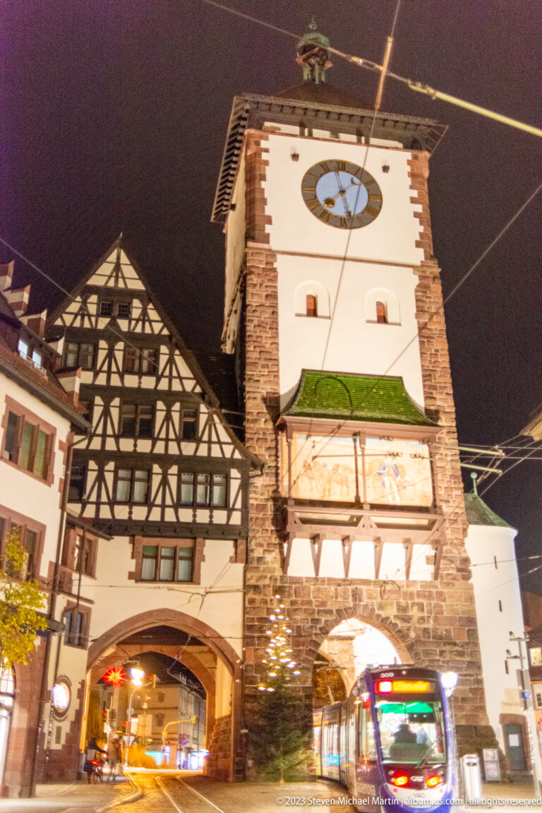 Illuminated Schwabentor with clock tower, painted panel, half-timbered wing, arched passage, and tram wires at night.