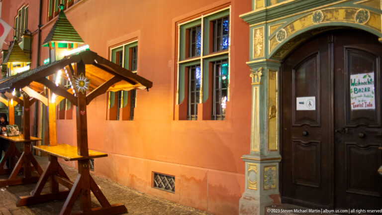 Small timber Christmas-market shelter and benches beside a warmly lit historic building and ornate doorway.