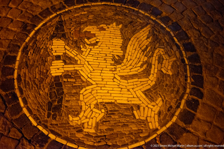 Circular cobblestone pavement mosaic depicting a stylized bird in contrasting light and dark stones.