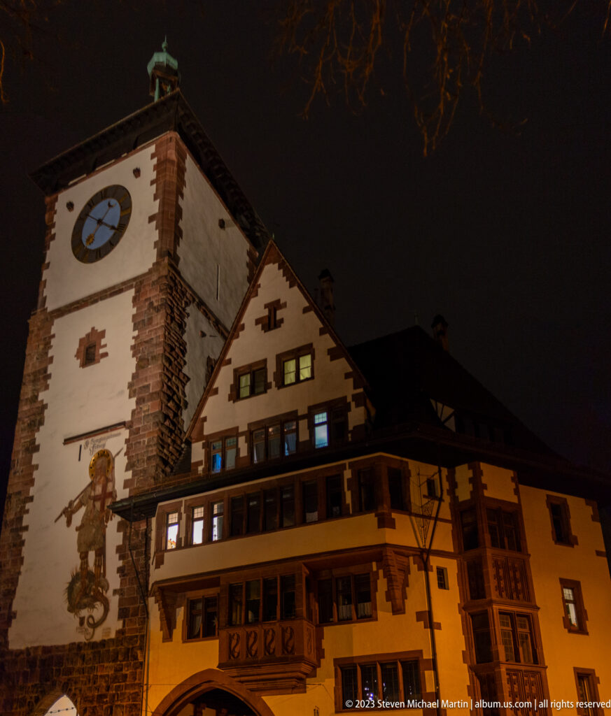 Illuminated historic gate tower with clock, painted wall figure, adjoining gabled building, and arched passage at night.