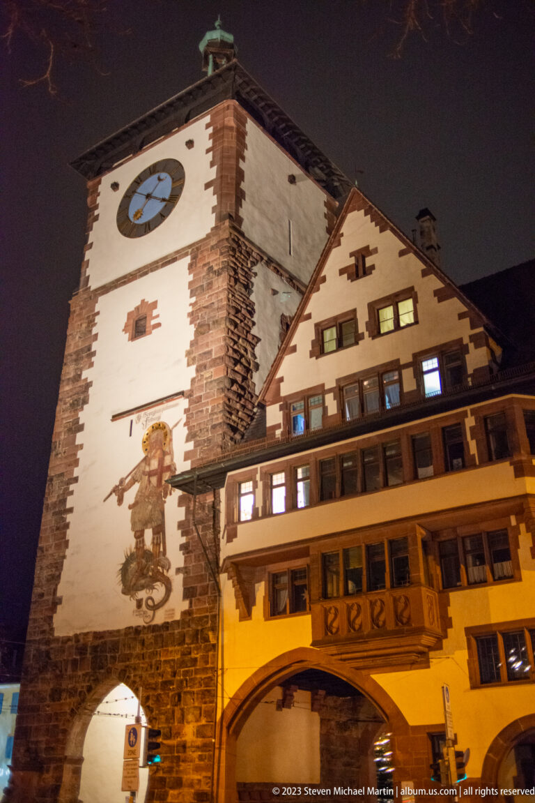 Closer night view of clocked gate tower beside a gabled building with balcony and lit windows.