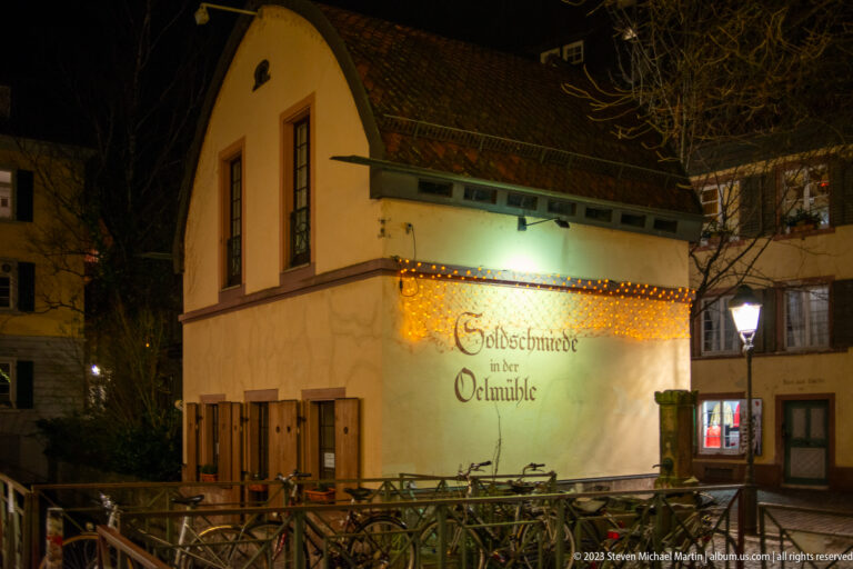 Small historic building at night with “Goldschmiede in der Ölmühle” lettering, wooden shutters, bicycles, and warm wall lighting.