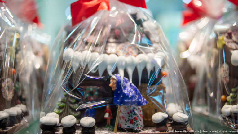 Close-up of clear glass Christmas ornament containing a small blue-and-white figurine, surrounded by blurred festive decorations.