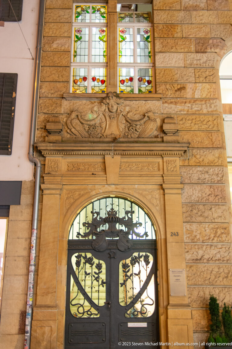 Ornate arched doorway with carved stone surround, wrought-iron door glazing, and stained-glass windows above.
