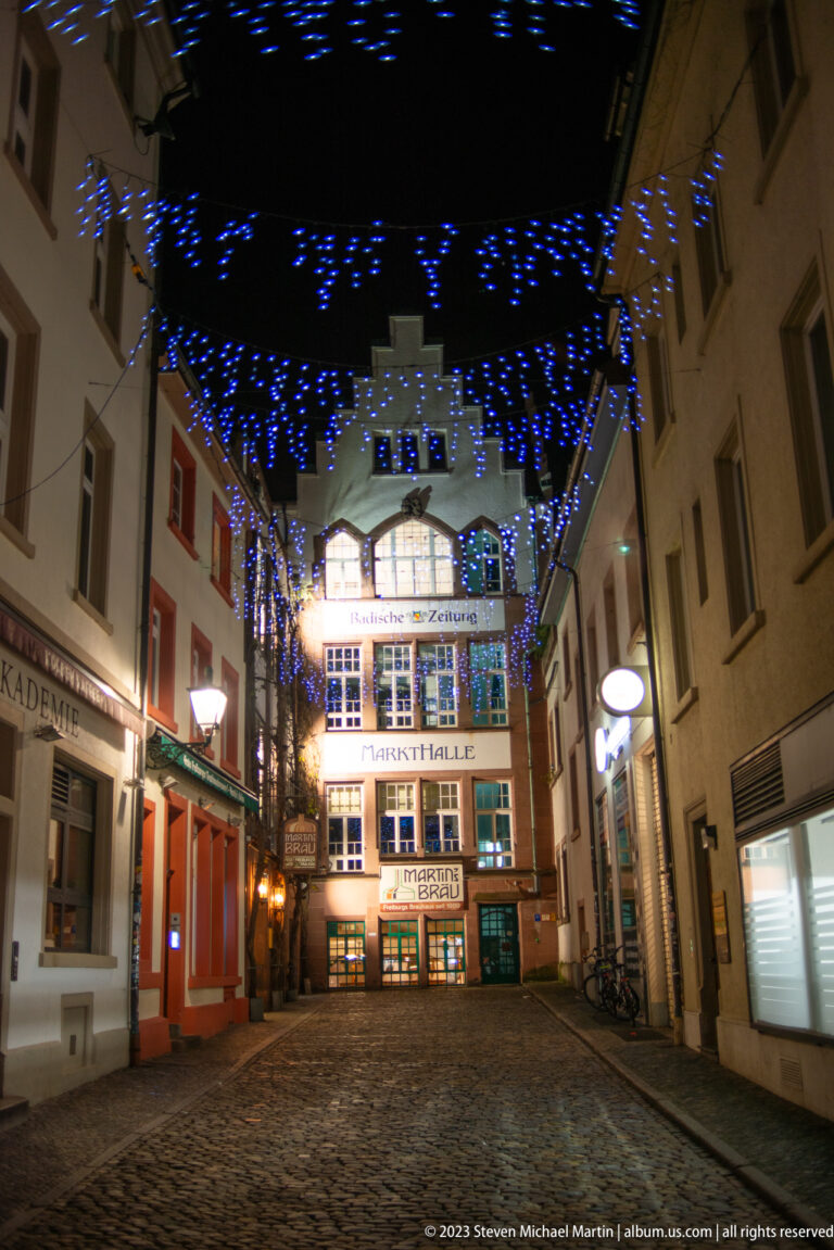 Narrow cobbled Freiburg street at night beneath blue-white Christmas lights, with shops and an illuminated historic building ahead.
