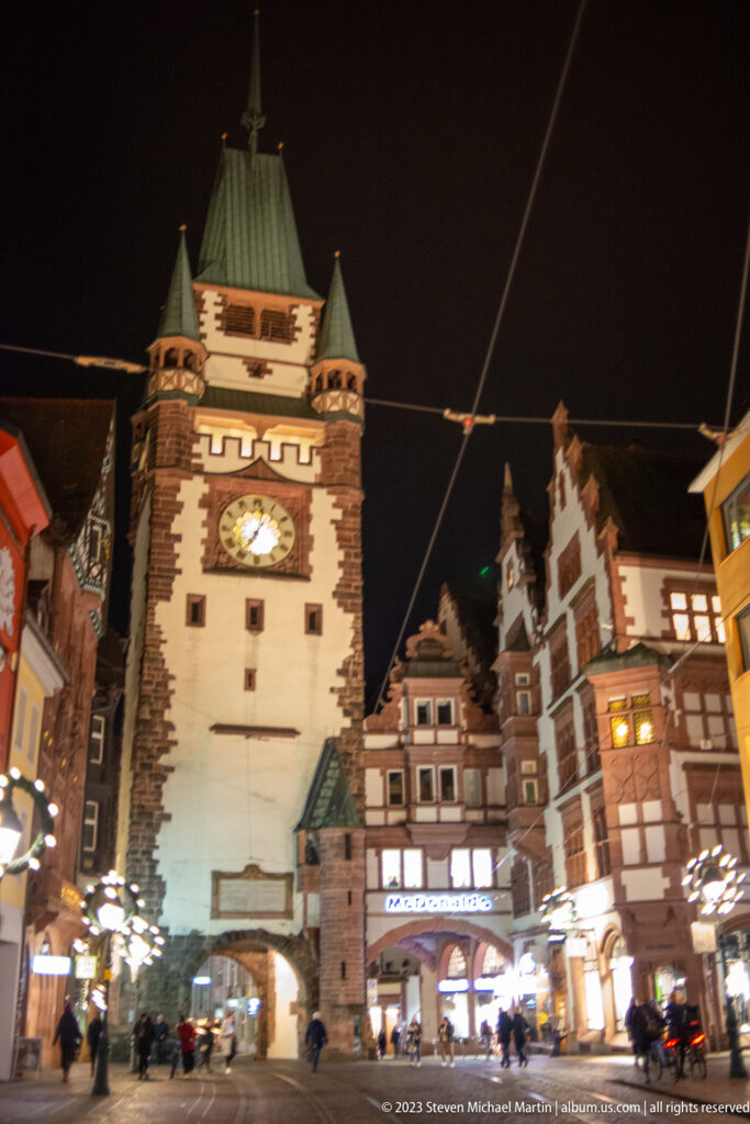 Martinstor at night with clock, pointed gateway, steep roof, tram wires, shops, and pedestrians in the surrounding street.