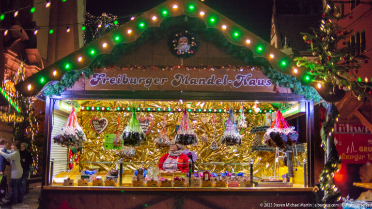 Illuminated Freiburger Mandel-Haus stall with vendor, hanging sweets, evergreen trim, and colorful Christmas lights.