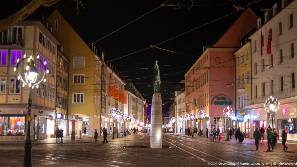 Bertoldsbrunnen column centered on a lit shopping street with tram tracks, overhead wires, pedestrians, and Christmas decorations.