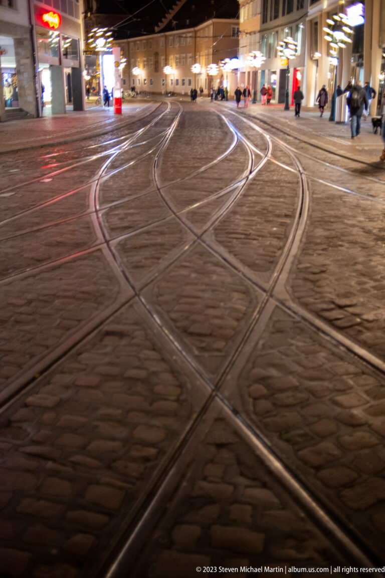 Curving and intersecting tram rails crossing cobblestones in Freiburg city center at night, with blurred lights beyond.