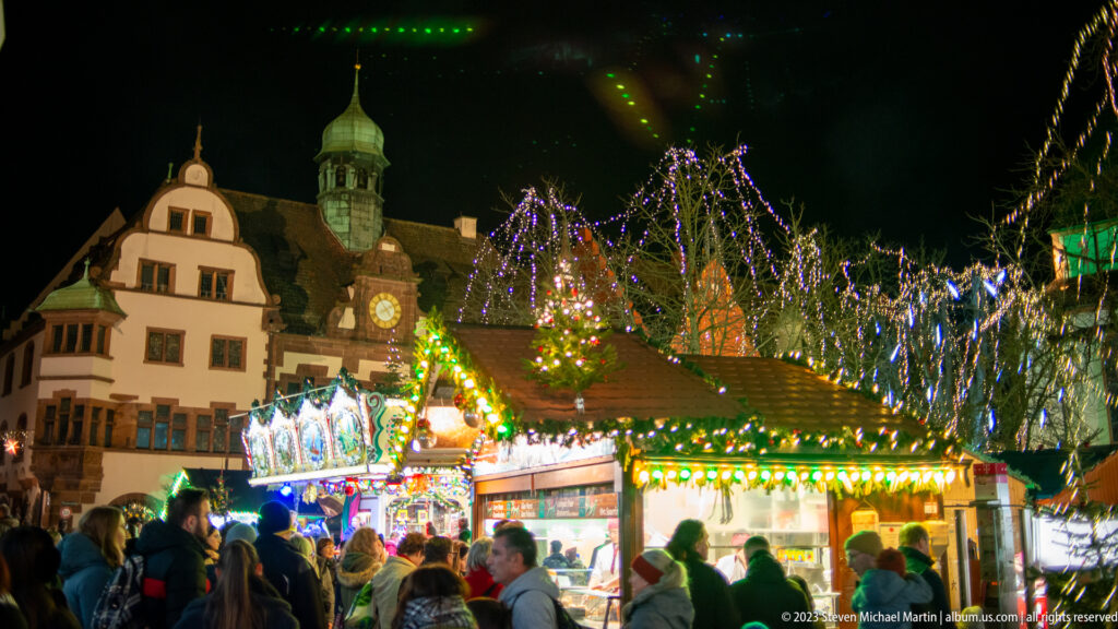 Crowded Freiburg Christmas market stall glowing with green lights, with historic buildings, a clock tower, and lit trees behind.