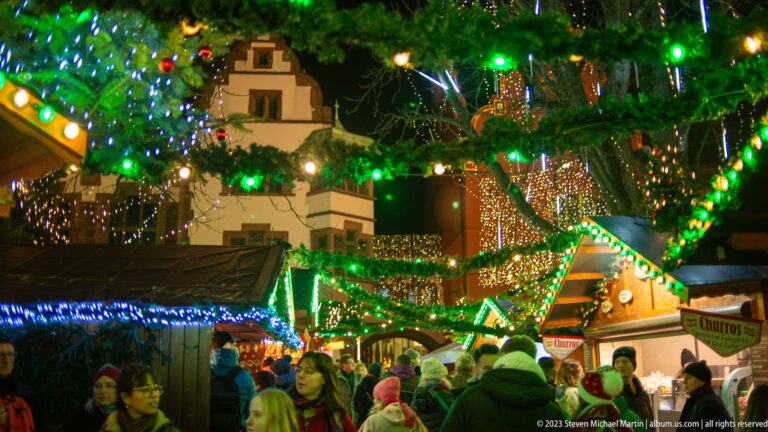 Crowded Christmas market lane beneath evergreen branches and multicolored lights, with decorated stalls and a visible churros sign.