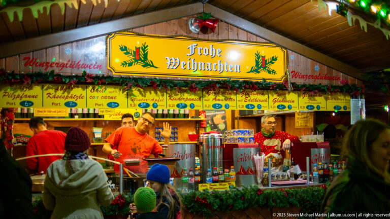 Christmas market food and drink stall with “Frohe Weihnachten” sign, vendors, garlands, and Glühwein signage.