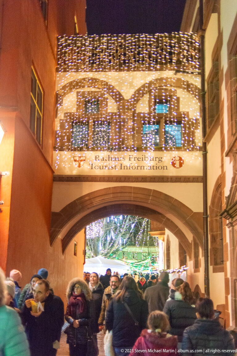 Large arch covered in warm-white Christmas lights above a crowd entering a more brightly decorated market area.