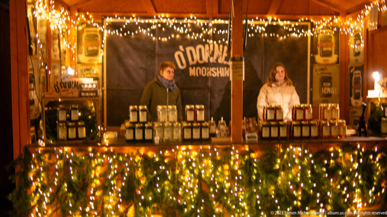 Warmly lit wooden Christmas market stall with two vendors behind rows of jars and bottles.
