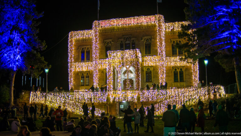 Historic Colombischlössle covered in warm-white Christmas lights, with blue-lit trees and a crowd gathered in front.